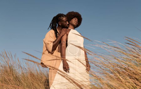 Foto de Vista lateral de la joven pareja afroamericana en vestidos de verano, mirando hacia otro lado mientras está de pie en el campo con hierba seca y macho con peinado de trenza afro abrazando a la mujer por detrás - Imagen libre de derechos