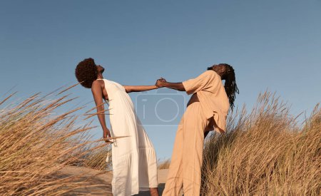 Foto de Vista lateral de ángulo bajo de la joven pareja afroamericana en vestidos de verano y peinado afro, mirando hacia arriba mientras está parado en el campo de arena de hierba seca cogido de la mano y apoyado contra el cielo azul sin nubes - Imagen libre de derechos