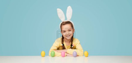 Foto de Niña feliz vistiendo orejas de conejito blanco mientras posa en la mesa blanca con una fila de huevos de colores en la parte superior - Imagen libre de derechos