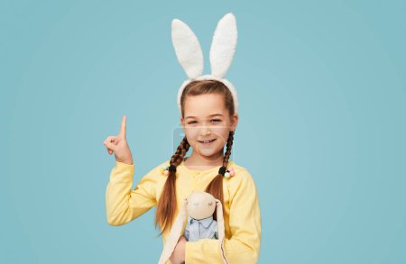 Foto de Encantadora chica de contenido con orejas de conejo de pie con juguete sobre fondo azul y apuntando hacia arriba - Imagen libre de derechos