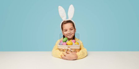Foto de Encantadora niña con orejas de conejo blanco y abrazando canasta de mimbre de huevos de Pascua de colores sobre fondo azul con espacio de copia - Imagen libre de derechos