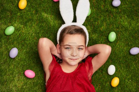 Téléchargez les photos : D'en haut de charmante fille portant des oreilles de lapin moelleux et couché de rêve sur l'herbe verte parmi les œufs de Pâques - en image libre de droit