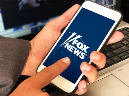 Téléchargez les photos : Man using fox news mobile application by touching on the screen with laptop in the backdrop. News editorial background - en image libre de droit
