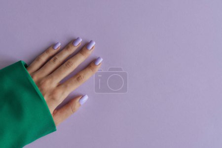 Gelische Maniküre mit violetten Nägeln auf lavendelfarbenem Hintergrund. Lila Fingernägel und grüne Kleidung
