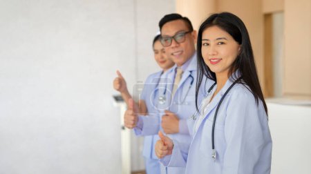 Téléchargez les photos : Rangée des médecins avec assistant travail d'équipe en uniforme, souriant, faire pouce vers le haut et debout devant un hôpital avec espace de copie pour le texte - en image libre de droit
