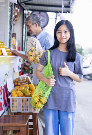 Porträt Teenie-Mädchen tragen Netztasche mit Orangen und tun Daumen nach oben vor der Gemeinde Obstladen für die Verwendung umweltfreundlicher Taschen, Hintergrund verschwommen ihr Großvater Orangen kaufen