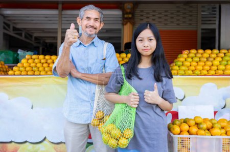 Porträt Teenagermädchen und älterer Großvater tragen Netztasche mit Orangen und tun Daumen hoch vor dem Obstladen der Gemeinde für die Unterstützung mit umweltfreundlichen Taschen