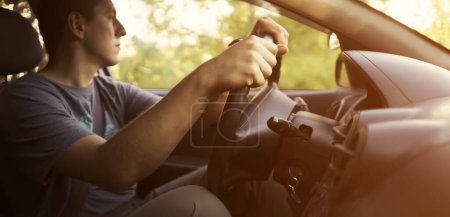 Téléchargez les photos : Un jeune homme voyage dans sa voiture, conduit le long de la route au coucher du soleil, tient le volant avec ses mains, vue rapprochée. - en image libre de droit