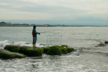 Foto de Sanur Beach, Bali, Indonesia (20 de febrero de 2021): La atmósfera de la playa de Sanur por la mañana, llena de olas y rocas, y un hombre pescando - Imagen libre de derechos