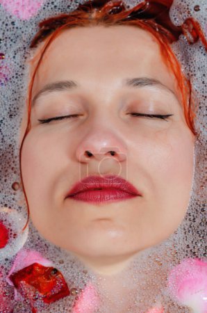 Foto de La cara de una hermosa mujer en el baño entre la espuma y pétalos de rosa - Imagen libre de derechos