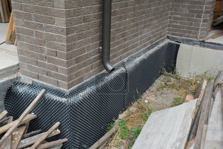 Fondation de maison sous-sol imperméabilisation, étanchéité humide avec isolation rigide. Installation d'une membrane d'étanchéité à gradins sur le mur extérieur du sous-sol et d'un tuyau de vidange de gouttière. 