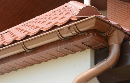 House metal rooftop with plastic rain gutter pipeline, soffit and soffit boards. Guttering house roof corner with downspout pipe in problem areas.