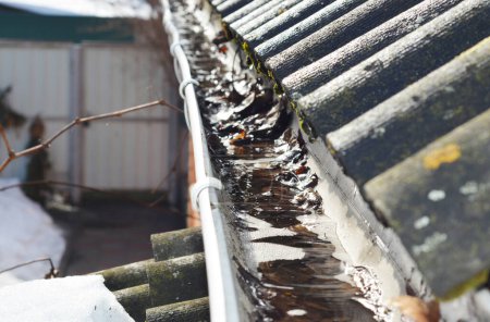 Photo for Spring roof gutter cleaning. A clogged rain gutter of an asbestos roof with non flowing water from melted snow because of unclean gutters with dirt and fallen leaves. - Royalty Free Image