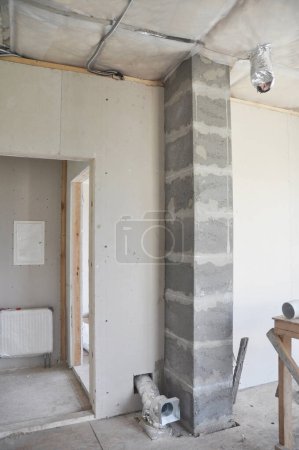 Ein Blick von innen auf den Rohbau eines Hauses mit einem Schornstein, der in einen Schornstein eingebaut wird, Lüftungsrohren, elektrischen Leitungen und Deckenisolierung. 