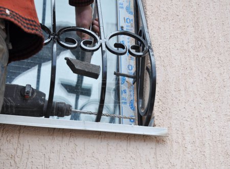 Foto de KYIV, UCRANIA - FEBRERO, 02, 2020: Protector de ventanas, instalación de parrilla. Un trabajador está instalando barras de seguridad de ventanas, barras antirrobo usando soportes de montaje interior, un martillo, un taladro eléctrico y un ancla. - Imagen libre de derechos