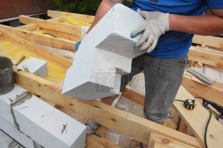 A building contractor installing an autoclaved aerated concrete block of irregular form, cut by a handsaw to lay it on the edge of a brick wall of the house construction where roof trusses installed.