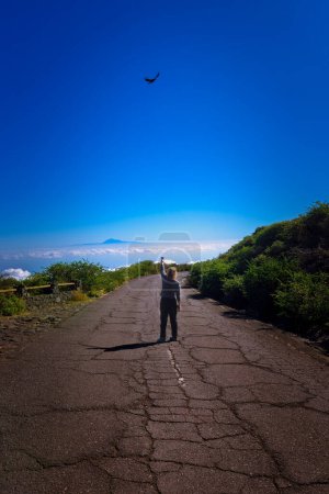 Unrecognizable woman with her back turned, in the middle of a high mountain road with a sea of clouds and the silhouette of a volcano in the background; pointing to a crow in flight.