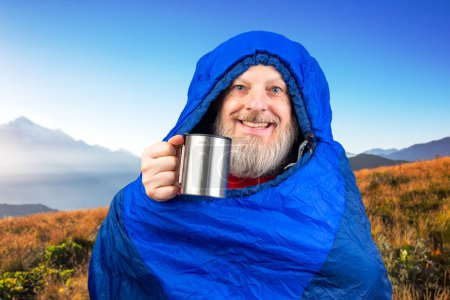 Glücklicher bärtiger Mann im Schlafsack mit einer Tasse Kaffee vor der Kulisse der Natur in den Bergen. Naturwanderungen in den Bergen