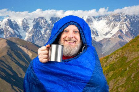 Glücklicher bärtiger Mann im Schlafsack mit einer Tasse Kaffee vor der Kulisse der Natur in den Bergen. Naturwanderungen in den Bergen