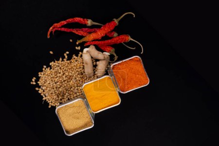 Photo for Three Square Bowls Filed With Spices. Isolated On Black Background. Spices Like, Coriander Powder, Red Chili Powder And Turmeric Powder. Coriander Seeds, Dried Red Chilies, Coriander Seeds And Raw Turmeric On Black Surface. - Royalty Free Image