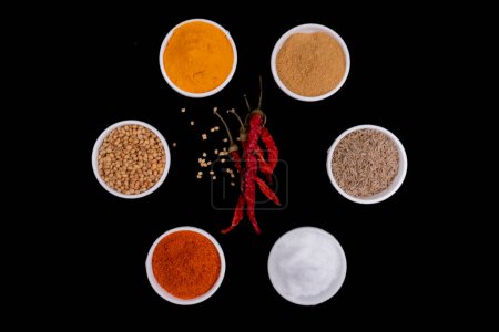 Photo for Six Round Bowls In Round Style Filled With Spices, In Center Of Dried Red Chilies With Seeds. Isolated On Black Background. Spices Like, Mustard Seeds, Coriander Seeds, Cumin Seeds, Red Chili Powder, Turmeric Powder And Salt. - Royalty Free Image