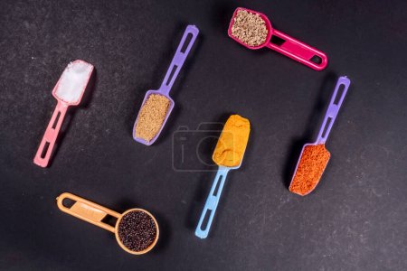 Photo for Multi Color Plastic Spoons In Spices Like Salt, Coriander Powder, Turmeric Powder, Red Chili Powder, Mustard Seeds And Ajwain On Black Paper Surface. Isolated On Black Paper Background. Copy Space - Royalty Free Image