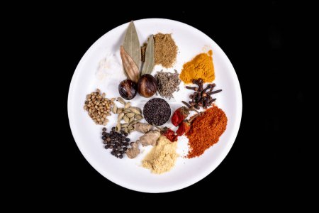 Photo for Top View Of White Dish Filed With Many Spices Like Spices Like, Mustard Seeds, Coriander Seeds, Cumin Seeds, Red Chili Powder, Turmeric Powder, Salt, Black Pepper, Clove, Cardamom Or Elaichi, Hing, Raw Turmeric And Nutmeg. Isolated On Black Backgroun - Royalty Free Image