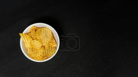 Téléchargez les photos : Chips de pomme de terre faits maison ou galette de pomme de terre dans un bol blanc, isolé sur fond de papier noir avec Copyspace - en image libre de droit
