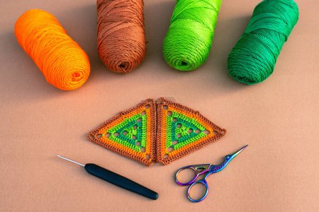 Photo for Two multicolored crochet motifs in green and orange tones, four skeins of yarn, crochet hook and small scissors on beige background. Concept of crochet supplies. - Royalty Free Image