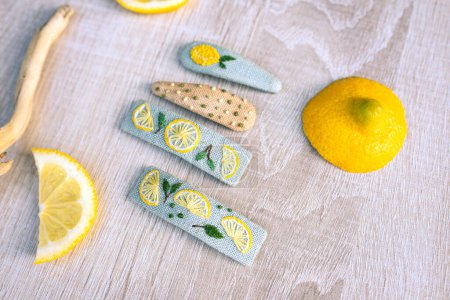 Photo for Snap hair clips with embroidered lemons and dots on them on white wooden background with slices of lemon around. Concept of creative embriodery. - Royalty Free Image