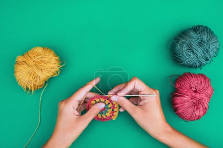 Close view of female hands, making a  pattern with crochet hook on green background with multicolored balls of yarn near. Crocheting a round motif in three colors.