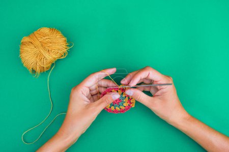 Téléchargez les photos : Vue des mains, faisant un motif rond au crochet sur fond vert avec un crochet et un fil jaune provenant de la boule de fil au motif. Vue simple et minimaliste sur l'artisanat. - en image libre de droit