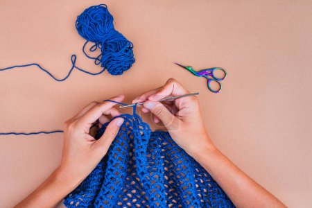 Vista de las manos tejiendo una red azul en backgorund beige con una madeja azul de hilo y tijeras azul-violeta arriba. Vista simple y minimalista sobre ganchillo.