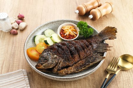 Photo for Gurame Bakar Madu, Grilled Gourami with honey and soy sauce. Served with sambal (chilli sauce), cucumber and tomatoes in white plate. - Royalty Free Image