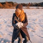 A woman in winter clothes collects a lump of snow in a snow-covered field.