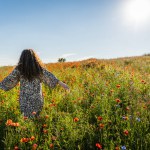 A curly long-haired plus size woman stands on a summer day among a poppy field with her arms spread to the sides.