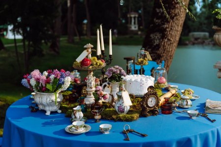 Der Tisch ist elegant dekoriert für eine Teeparty im Stil von Alice im Wunderland. Tassen und Untertassen, Kerzen im Kerzenständer, Kuchen, alte Schlüssel, Blumen in der Vase, eine Uhr auf einer blauen Tischdecke spät