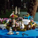 The table is elegantly decorated for a tea party in the style of Alice in Wonderland. Cups and saucers, candles in a candlestick, cake, old keys, flowers in a vase, a clock on a blue tablecloth late