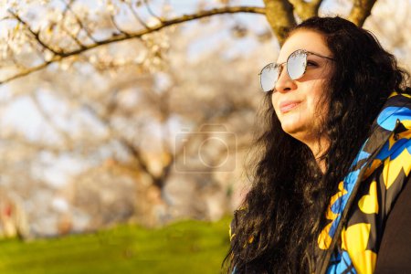 Woman in sunglasses with mirrored lenses with long dark hair in blooming spring garden in public park on sunny spring day.