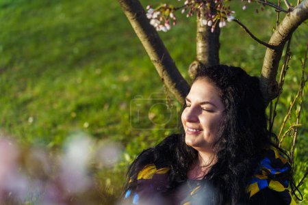 Young woman sitting in blooming cherry orchard leaning on tree trunk with closed eyes and enjoying spring sunny day.