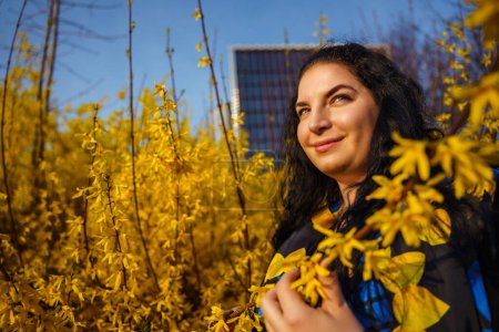 Young happy smiling woman plus size in spring in city park among blooming yellow forsythia bushes.