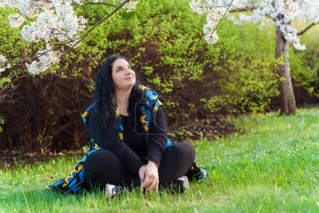 Young plus size woman in blue and yellow vest admiring the beauty of cherry blossom trees in the park in spring.