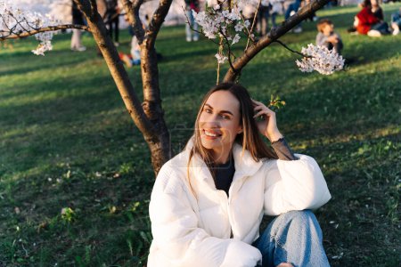Smiling cheerful woman in white down jacket and jeans sitting on grass in city park in spring under cherry blossom.