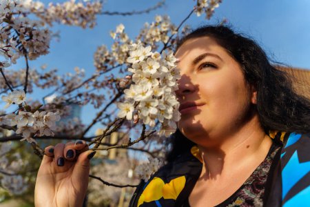 Atractiva mujer joven de tamaño grande oliendo rama de flor de cerezo en el parque en primavera.