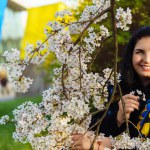 Long-haired young Ukrainian woman brunette getting pleasure and smiling while spending time among flowering trees in city park.