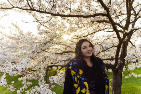 Young long-haired plus size woman spending time among cherry blossom trees in city park in spring.