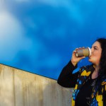 Beautiful young brunette woman with long curly hair, dressed in a black, blue and yellow waistcoat, drinks coffee from a paper cup on a background of blue wall.