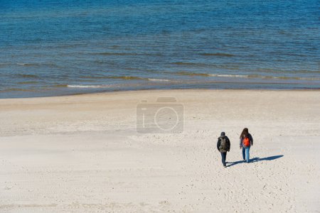 Photo for A couple of travelers in warm clothes with backpacks walk along a deserted beach near the sea in the off-season. Curonian Spit, Neringa, Lithuania. - Royalty Free Image