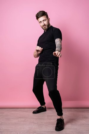 Téléchargez les photos : Pleine grandeur de beau, drôle, fou, émotionnel, beau jeune homme barbu portant en total noir vêtements drôle danse isolé sur fond rose - en image libre de droit