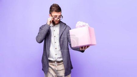 Photo for Handsome bearded man looks skeptically at a pink gift box holding it in his hand and adjusting his glasses isolated on purple background - Royalty Free Image